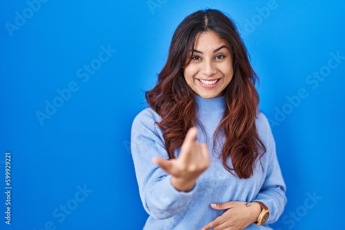 Hispanic young woman standing over blue background beckoning come here gesture with hand inviting welcoming happy and smiling