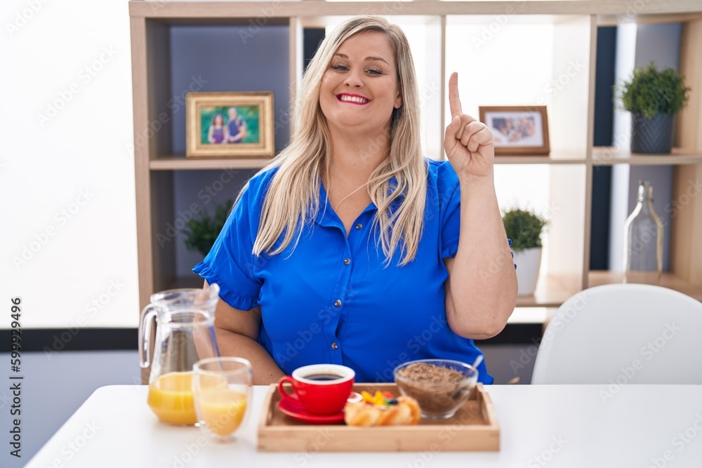 Caucasian plus size woman eating breakfast at home showing and pointing up with finger number one while smiling confident and happy. Caucasian plus size woman eating breakfast at home showing and pointing up with finger number one while smiling confident and happy.