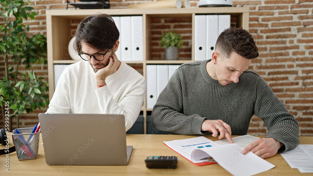 Two men business partners using laptop writing on document working at office Two men business partners using laptop writing on document working at office