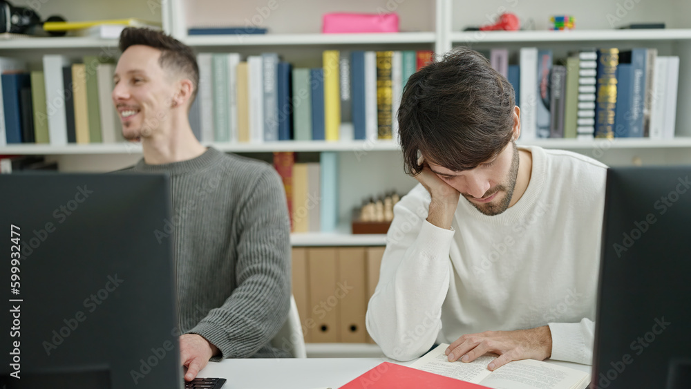 Two men students using computer reading book studying at library university Two men students using computer reading book studying at library university