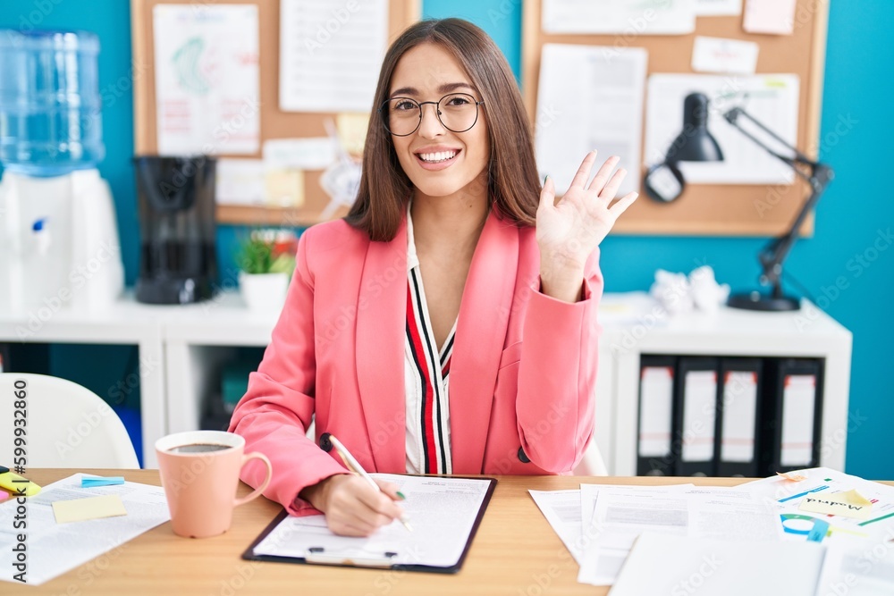 Young hispanic woman working at the office wearing glasses waiving saying hello happy and smiling, friendly welcome gesture