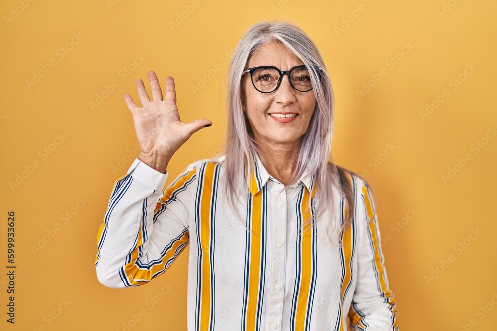 Φωτογραφία Middle age woman with grey hair standing over yellow background wearing glasses showing and pointing up with fingers number five while smiling confident and happy