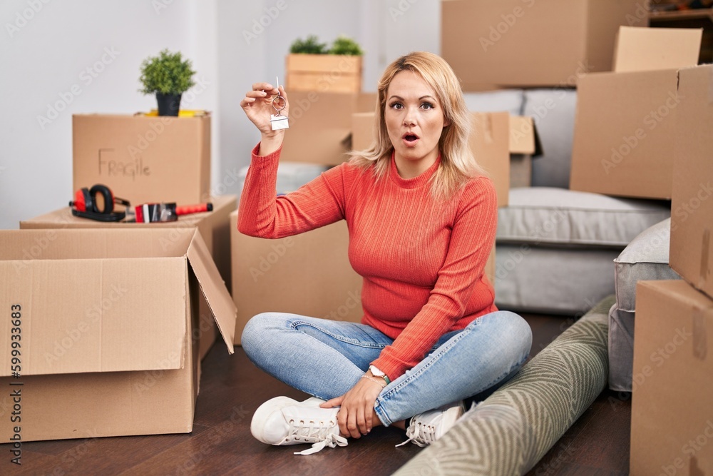 Blonde woman holding keys of new home sitting on the floor scared and amazed with open mouth for surprise, disbelief face Blonde woman holding keys of new home sitting on the floor scared and amazed with open mouth for surprise, disbelief face