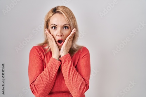Blonde woman standing over isolated background afraid and shocked, surprise and amazed expression with hands on face