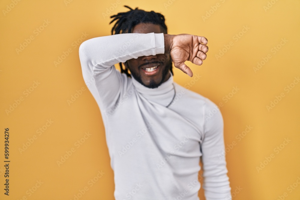 African man with dreadlocks wearing turtleneck sweater over yellow background covering eyes with arm smiling cheerful and funny. blind concept. African man with dreadlocks wearing turtleneck sweater over yellow background covering eyes with arm smiling cheerful and funny. blind concept.