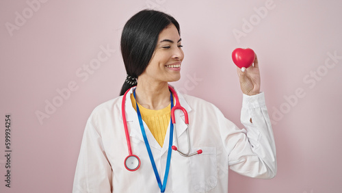 Young beautiful hispanic woman doctor smiling confident holding heart over isolated pink background