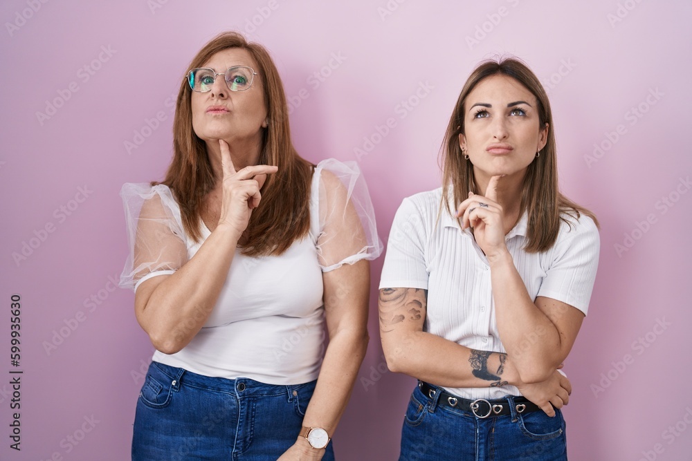 Hispanic mother and daughter wearing casual white t shirt over pink background thinking concentrated about doubt with finger on chin and looking up wondering Hispanic mother and daughter wearing casual white t shirt over pink background thinking concentrated about doubt with finger on chin and looking up wondering