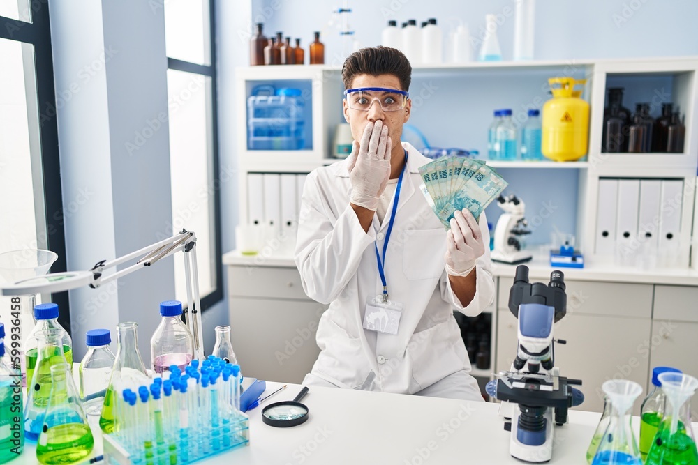 Young hispanic man working at scientist laboratory holding brazilian reals covering mouth with hand, shocked and afraid for mistake. surprised expression Young hispanic man working at scientist laboratory holding brazilian reals covering mouth with hand, shocked and afraid for mistake. surprised expression