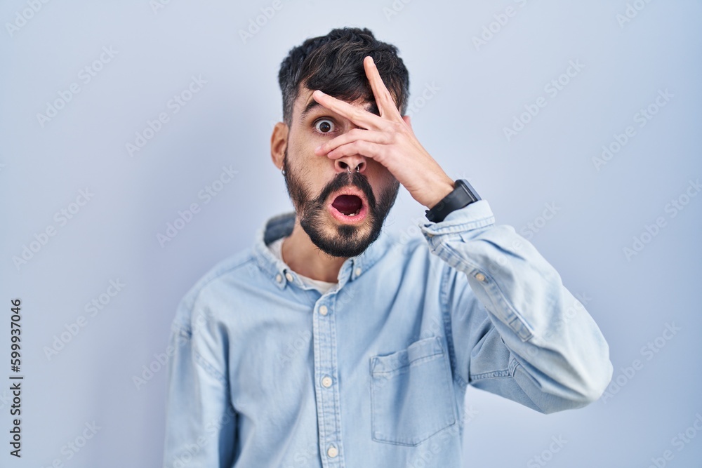 Young hispanic man with beard standing over blue background peeking in shock covering face and eyes with hand, looking through fingers with embarrassed expression. Young hispanic man with beard standing over blue background peeking in shock covering face and eyes with hand, looking through fingers with embarrassed expression.