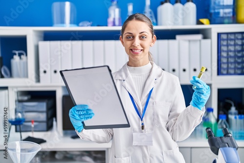Young woman scientist smiling confident reading document holding test tube at laboratory