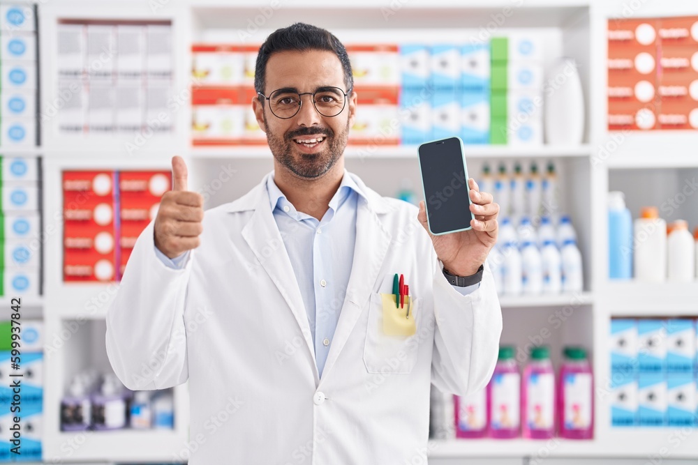 Hispanic man with beard working at pharmacy drugstore showing smartphone screen smiling happy and positive, thumb up doing excellent and approval sign