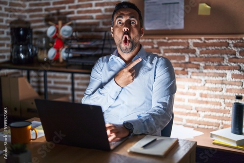 Hispanic man with beard working at the office at night surprised pointing with finger to the side, open mouth amazed expression.