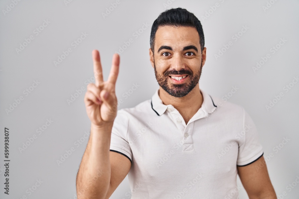 Fototapeta premium Young hispanic man with beard wearing casual clothes over white background showing and pointing up with fingers number two while smiling confident and happy.