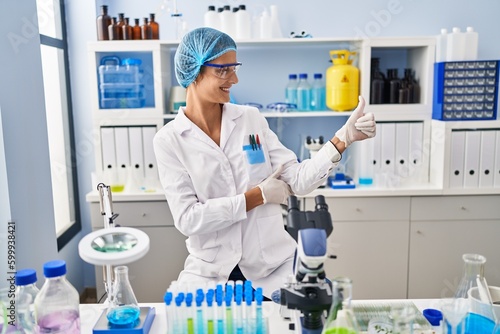 Brunette woman working at scientist laboratory looking proud  smiling doing thumbs up gesture to the side