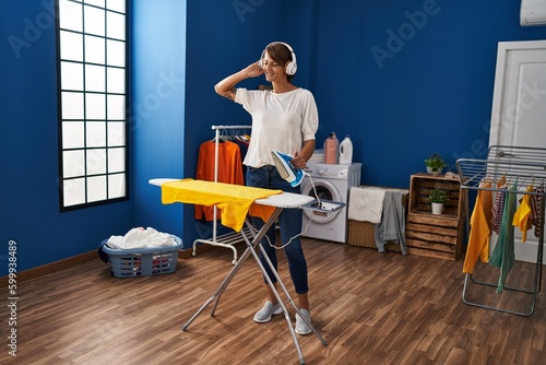 Young beautiful hispanic woman listening to music ironing clothes at laundry room