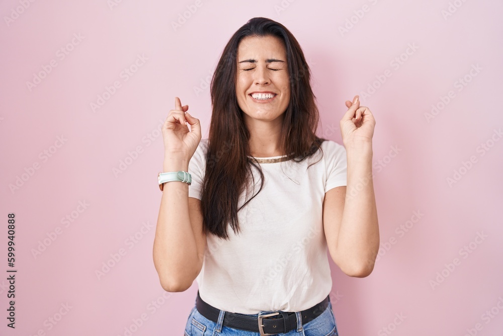 Young brunette woman standing over pink background gesturing finger crossed smiling with hope and eyes closed. luck and superstitious concept. Young brunette woman standing over pink background gesturing finger crossed smiling with hope and eyes closed. luck and superstitious concept.