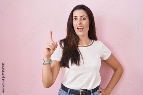Young brunette woman standing over pink background pointing finger up with successful idea. exited and happy. number one.
