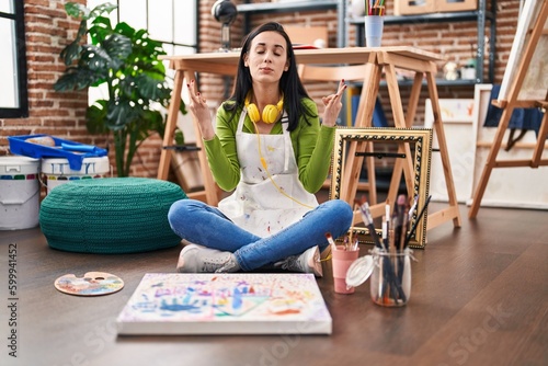Hispanic woman sitting at art studio painting on canvas relax and smiling with eyes closed doing meditation gesture with fingers. yoga concept.
