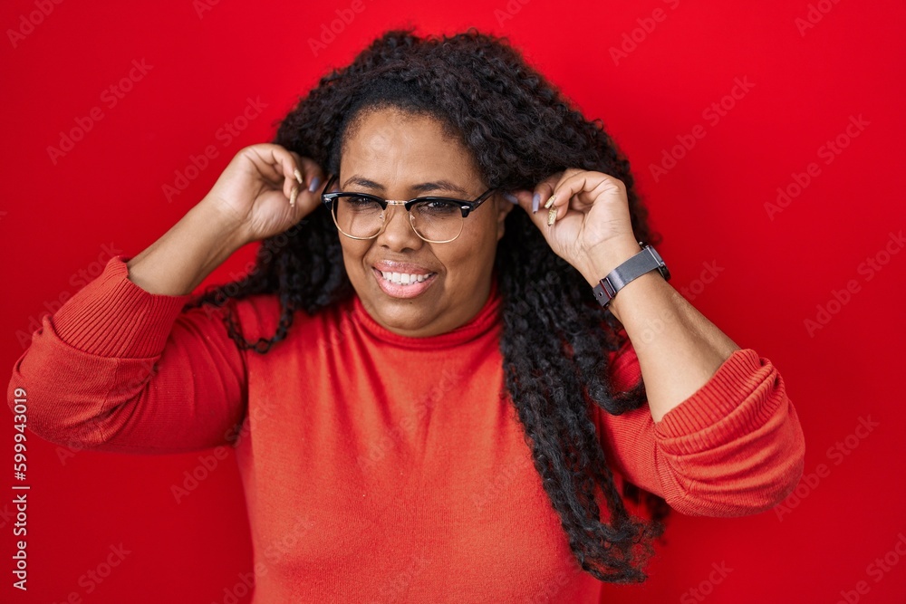 Plus size hispanic woman standing over red background smiling pulling ears with fingers, funny gesture. audition problem Plus size hispanic woman standing over red background smiling pulling ears with fingers, funny gesture. audition problem