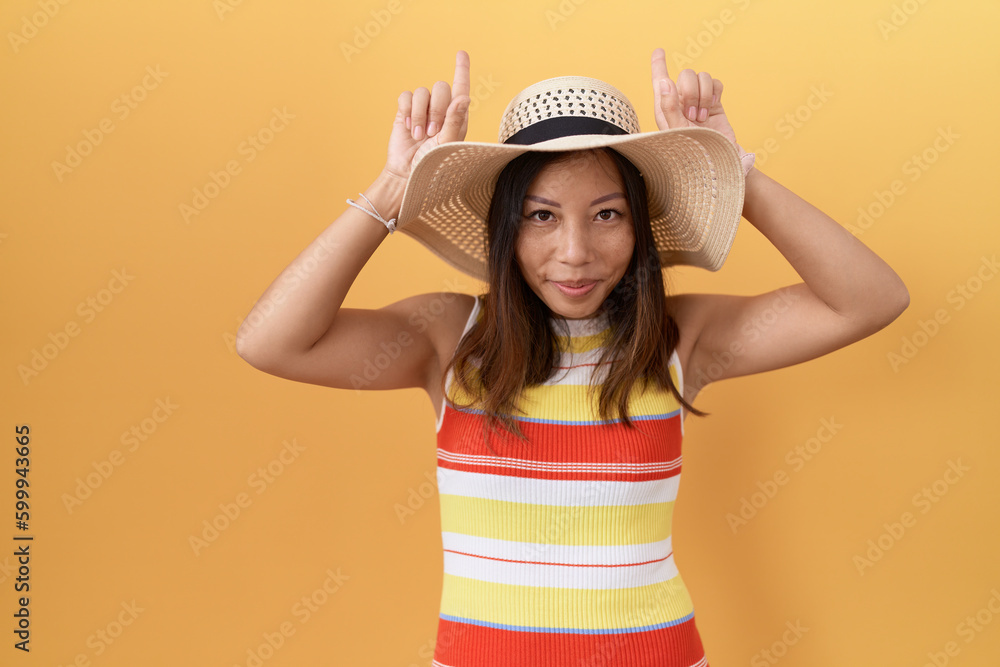 Middle age chinese woman wearing summer hat over yellow background doing funny gesture with finger over head as bull horns Middle age chinese woman wearing summer hat over yellow background doing funny gesture with finger over head as bull horns