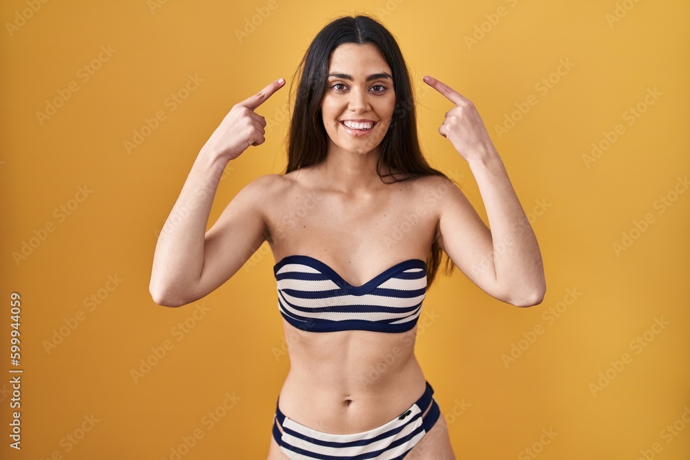 Young brunette woman wearing bikini over yellow background smiling pointing to head with both hands finger, great idea or thought, good memory Young brunette woman wearing bikini over yellow background smiling pointing to head with both hands finger, great idea or thought, good memory
