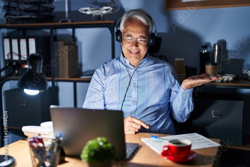 Hispanic senior man wearing call center agent headset at night smiling cheerful presenting and pointing with palm of hand looking at the camera.