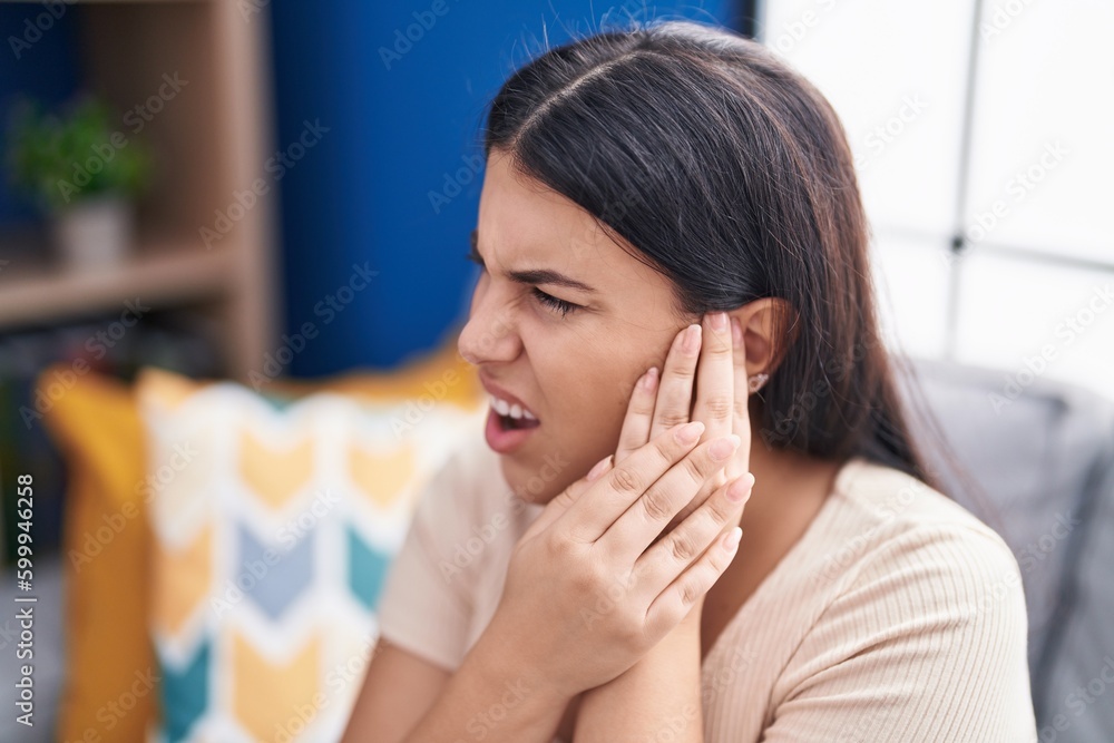 Young beautiful hispanic woman suffering for ear pain sitting on sofa at home Young beautiful hispanic woman suffering for ear pain sitting on sofa at home