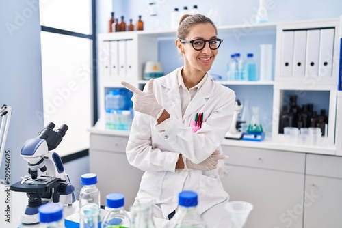 Young woman working at scientist laboratory smiling cheerful pointing with hand and finger up to the side