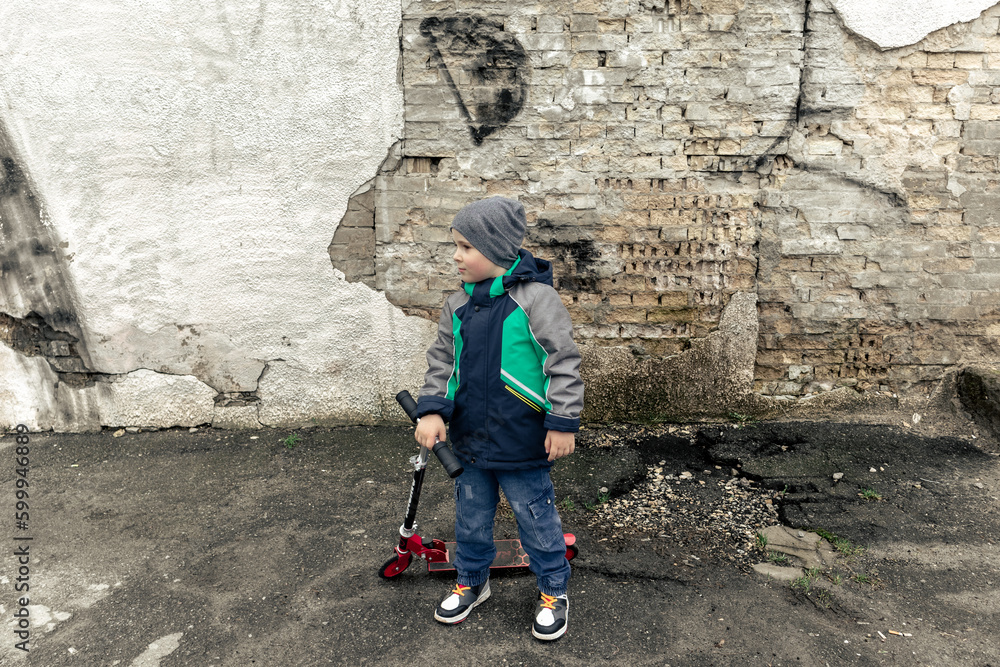 A little boy with a scooter on the background of a broken brick wall