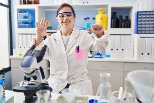 Hispanic girl with down syndrome working at scientist laboratory showing and pointing up with fingers number six while smiling confident and happy.
