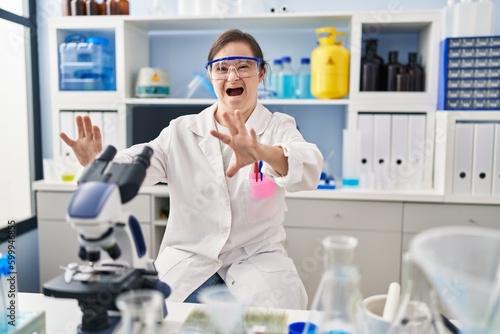 Hispanic girl with down syndrome working at scientist laboratory afraid and terrified with fear expression stop gesture with hands  shouting in shock. panic concept.