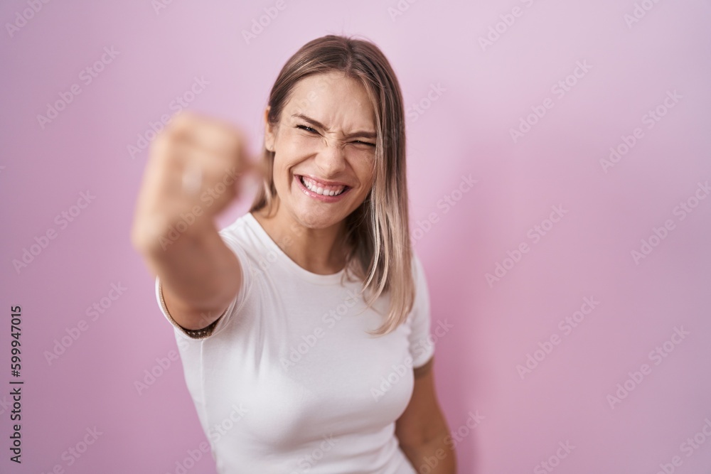 Blonde caucasian woman standing over pink background angry and mad raising fist frustrated and furious while shouting with anger. rage and aggressive concept. Blonde caucasian woman standing over pink background angry and mad raising fist frustrated and furious while shouting with anger. rage and aggressive concept.