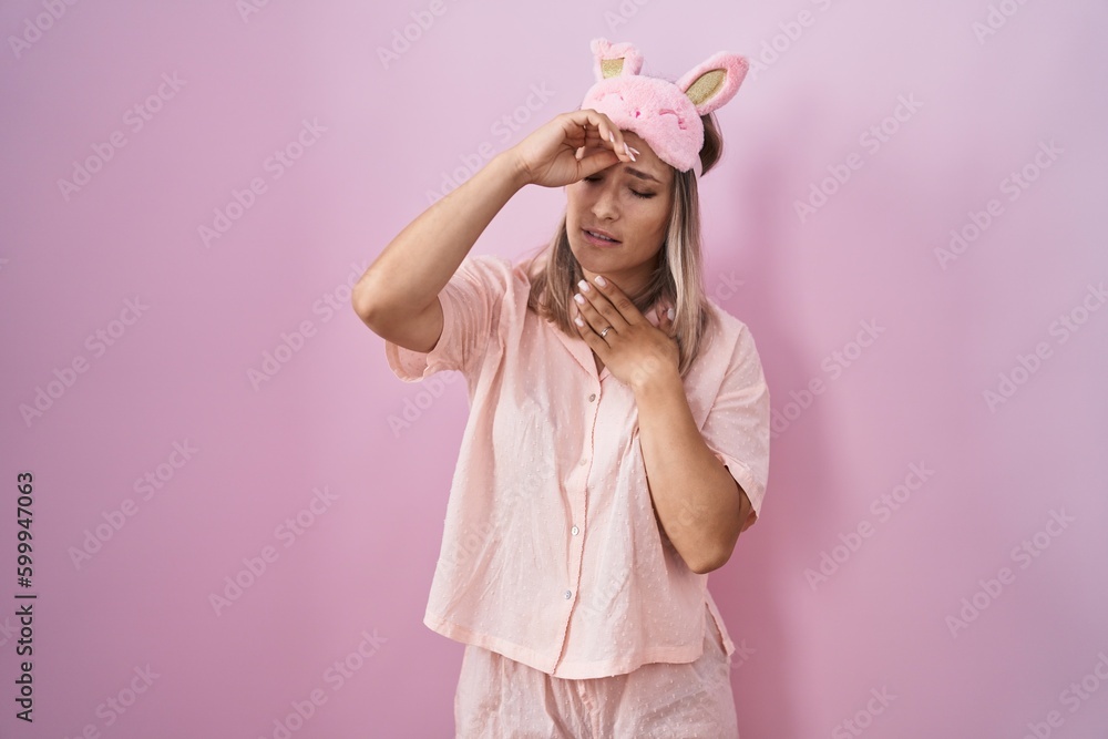 Blonde caucasian woman wearing sleep mask and pajama touching forehead for illness and fever, flu and cold, virus sick