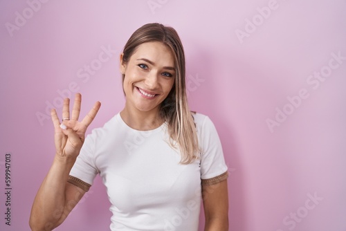 Blonde caucasian woman standing over pink background showing and pointing up with fingers number four while smiling confident and happy.