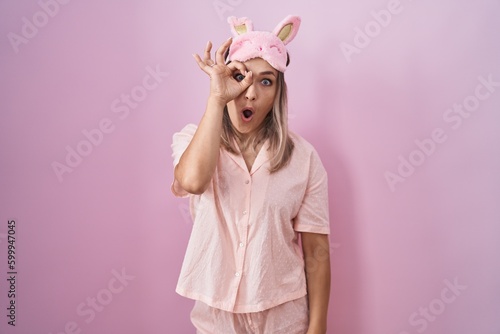 Blonde caucasian woman wearing sleep mask and pajama doing ok gesture shocked with surprised face, eye looking through fingers. unbelieving expression.