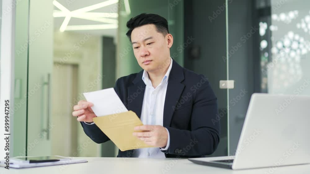 Disappointed asian man reading letter with bad news while sitting at desk at workplace in modern office. Worried sad businessman in formal suit frustrated by received unpleasant notification Disappointed asian man reading letter with bad news while sitting at desk at workplace in modern office. Worried sad businessman in formal suit frustrated by received unpleasant notification