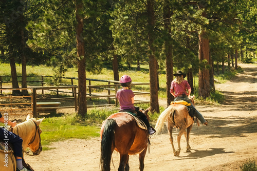 Young woman in wide-brimmed hat riding horse on trail in the woods while young girl and another person following her