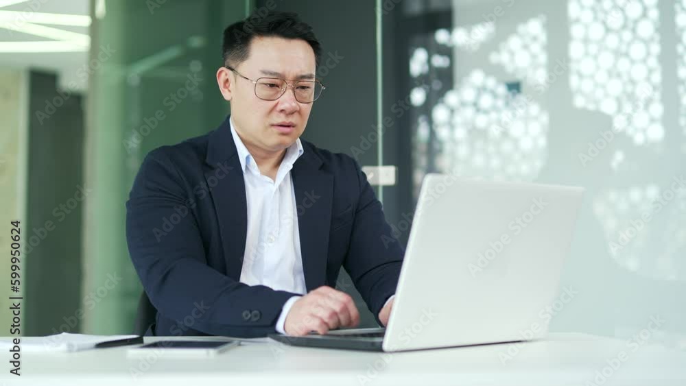 Overworked asian man suffering from eye strain while working on laptop while sitting at desk at workplace in modern office. Businessman takes off his glasses, massages and rubs the bridge of his nose Overworked asian man suffering from eye strain while working on laptop while sitting at desk at workplace in modern office. Businessman takes off his glasses, massages and rubs the bridge of his nose
