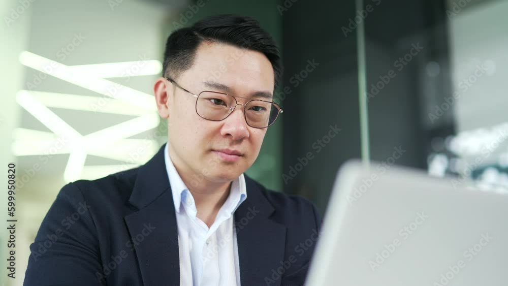 Close up of smiling asian businessman in formal suit working typing on laptop while sitting at desk at workplace in modern office. Happy entrepreneur looks at or writes a message to a client by email Close up of smiling asian businessman in formal suit working typing on laptop while sitting at desk at workplace in modern office. Happy entrepreneur looks at or writes a message to a client by email