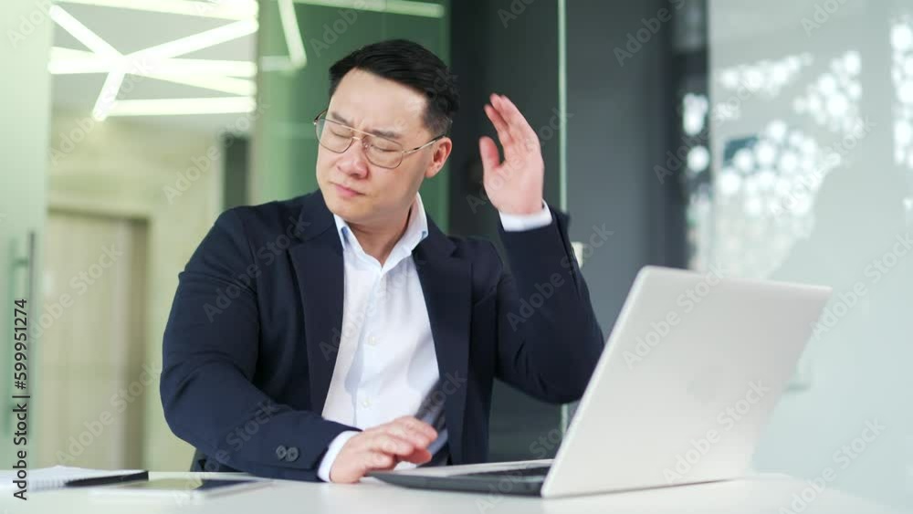Sick asian man suffering from neck pain while working on laptop while sitting at desk at workplace in modern office. Businessman in a formal suit and glasses stretches, massages and rubs sore muscles Sick asian man suffering from neck pain while working on laptop while sitting at desk at workplace in modern office. Businessman in a formal suit and glasses stretches, massages and rubs sore muscles