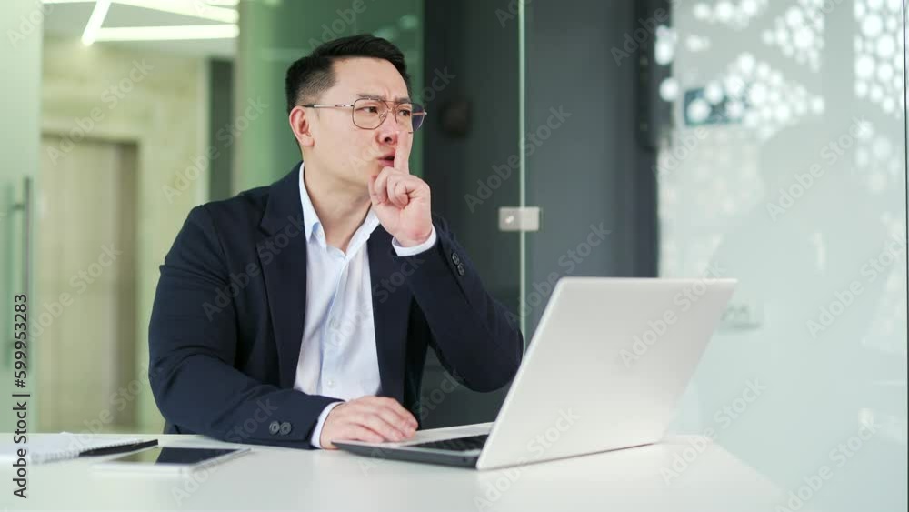 Asian man holding index finger to lips while working on laptop while sitting at workplace in office. Еmployee covers his ears, shows a gesture of silence. Says shh, asks not to make noise, to be quiet