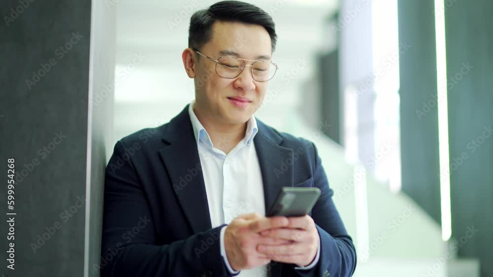 Smiling asian businessman in a formal suit and glasses is using a smartphone while standing in a modern office. Confident happy owner is browsing checking messages, chatting online or texting a client Smiling asian businessman in a formal suit and glasses is using a smartphone while standing in a modern office. Confident happy owner is browsing checking messages, chatting online or texting a client