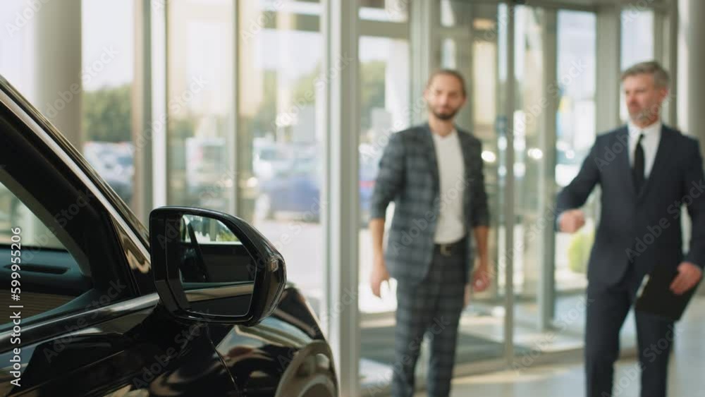 Young successful customer man deciding to buy new auto in showroom. Happy car salesman and male customer in auto dealership, they are shaking hands seal the purchase of the auto or new car Young successful customer man deciding to buy new auto in showroom. Happy car salesman and male customer in auto dealership, they are shaking hands seal the purchase of the auto or new car