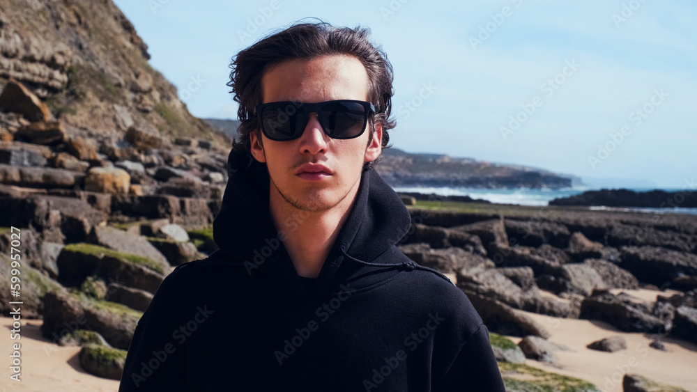 Portrait of an young man with brown hair blown on wind against rock on summer sunny day. Attractive young man looks at the camera on the nature. Handsome guy in black sunglasses poses outdoors. Portrait of an young man with brown hair blown on wind against rock on summer sunny day. Attractive young man looks at the camera on the nature. Handsome guy in black sunglasses poses outdoors.