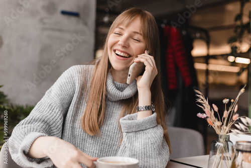 Blonde woman smiling positive conversation talking to her friend using phone hand hold hot drink tea relax casual positive emotion weekend relax routine at cafe restaurant. Girl have discussion.