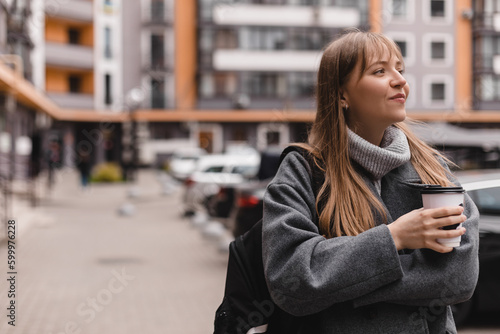 Cheerful young blonde woman with bang in casual wear drinking takeaway coffee while walking outdoors at street. Girl wear grey sweater, coat and black backpack. Woman walk or go home in courtyard. © zvkate
