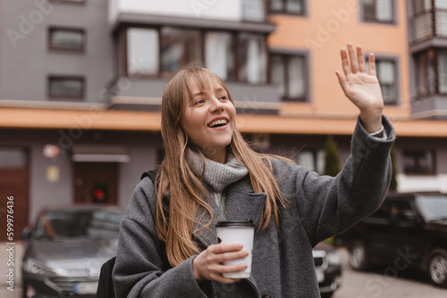 Smiling young woman waving her hand with a friendly cheerful smile to her neighbours. Girl wear grey sweater and coat, hold cup of tea, meeting friends, welcome, nice to meet you. Hi, hello gesture.