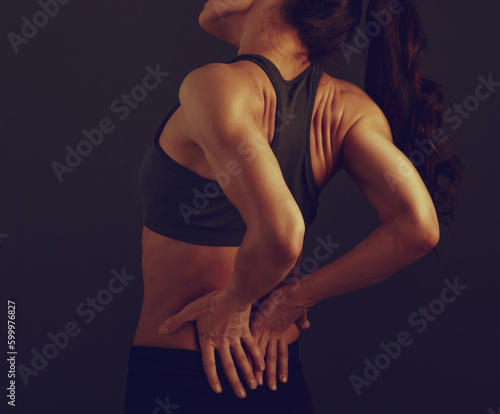 Female sporty muscular model with ponytail doing correction massage holding two hands on the low back on dark shadow grey background with empty copy space. photo