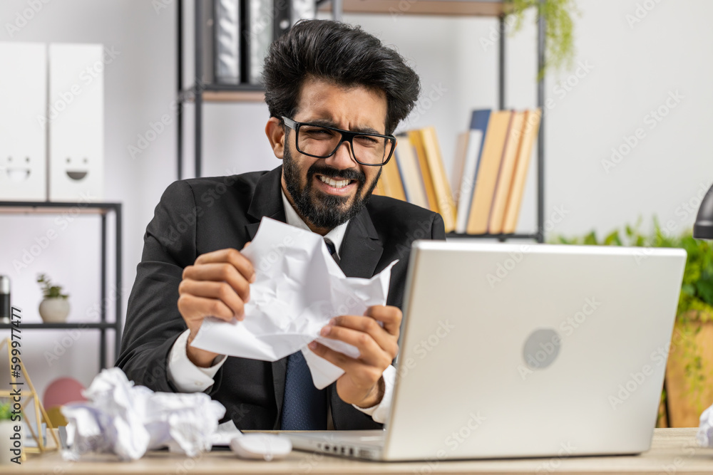 Angry furious indian man working at office throwing crumpled paper, having nervous breakdown at work, migaine, headache, stress management, mental distress problems, losing temper, reaction on failure Angry furious indian man working at office throwing crumpled paper, having nervous breakdown at work, migaine, headache, stress management, mental distress problems, losing temper, reaction on failure