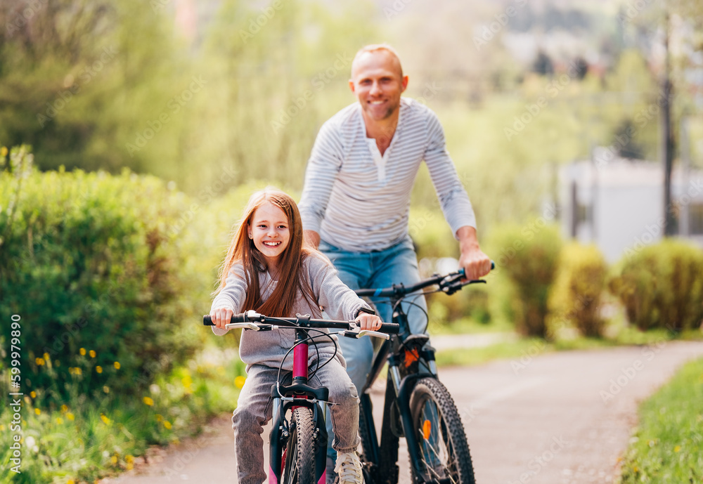 Portraits Smiling father with daughter during summer outdoor bicycle riding. They enjoy togetherness in the summer city park. Happy parenthood and childhood or active sport life concept image.